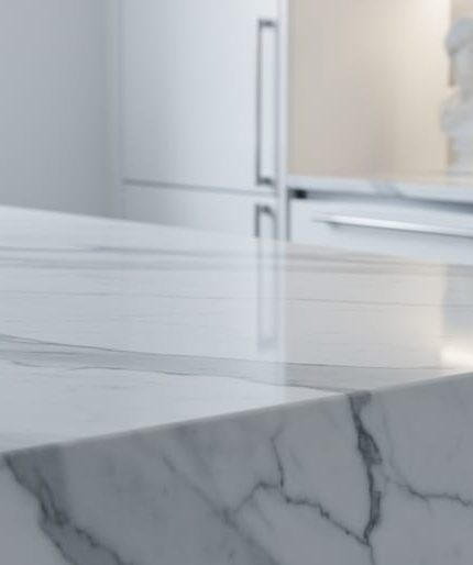Close-up of a polished white Carrara marble kitchen countertop with gray veining, side-lit to show subtle translucency, with a softly blurred modern kitchen and a small classical marble bust in the background.