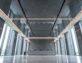 Low-angle view of a modern lobby showcasing a polished granite ceiling with coffered panels, reveal joints, and recessed LED lighting, with a minimal reception desk and stone walls fading into the background.