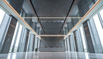 Low-angle view of a modern lobby showcasing a polished granite ceiling with coffered panels, reveal joints, and recessed LED lighting, with a minimal reception desk and stone walls fading into the background.
