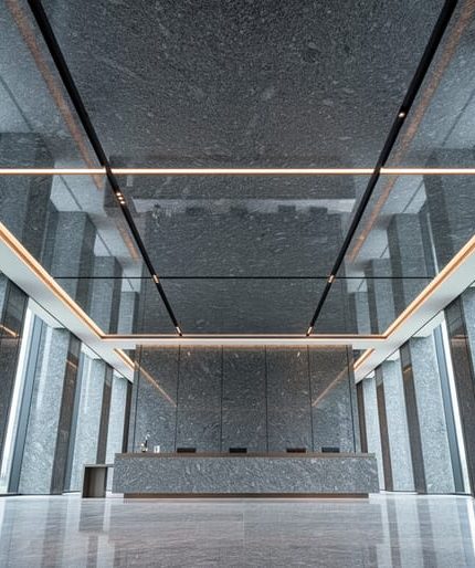 Low-angle view of a modern lobby showcasing a polished granite ceiling with coffered panels, reveal joints, and recessed LED lighting, with a minimal reception desk and stone walls fading into the background.