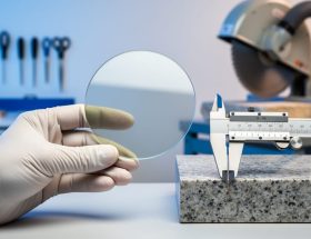 Gloved hand holding a reflective silicon wafer next to a polished granite sample and calipers on a clean bench, with a softly blurred background showing cleanroom tools on one side and a stone-cutting saw with dust extraction on the other.