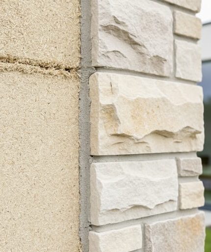 Close-up of a modern sustainable building wall where a pale hemp-lime (hempcrete) surface meets a limestone stone exterior, showing a detailed material junction in natural light.