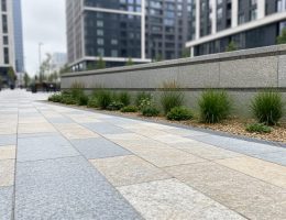 Eye-level view of a modern urban plaza with natural stone pavers, a low retaining wall, and integrated greenery.