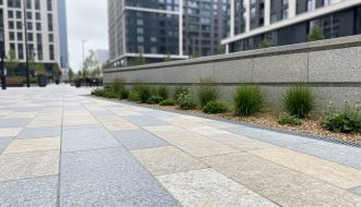 Eye-level view of a modern urban plaza with natural stone pavers, a low retaining wall, and integrated greenery.